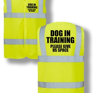 Op de afbeelding: Gele veiligheidsvest met reflecterende strepen en de tekst "DOG IN TRAINING PLEASE GIVE US SPACE" in het zwart gedrukt.
