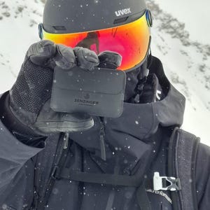 Könnte beinhalten: Eine schwarze Geldbörse mit dem Text "Zenzinger" wird vor einer Person gehalten, die einen schwarzen Helm mit einem reflektierenden orangefarbenen Visier und schwarzen Handschuhen trägt. Die Person trägt eine schwarze Jacke in einer verschneiten Umgebung.