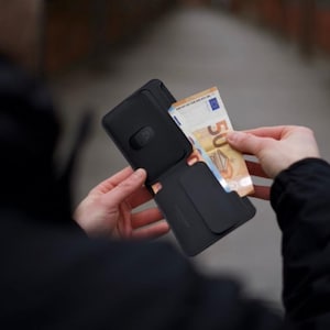Könnte beinhalten: Eine schwarze Geldbörse ist geöffnet und zeigt einen Fünfzig-Euro-Schein. Die Geldbörse wird von den Händen einer Person offen gehalten. Die Geldbörse hat ein minimalistisches Design mit Magnetverschluss.