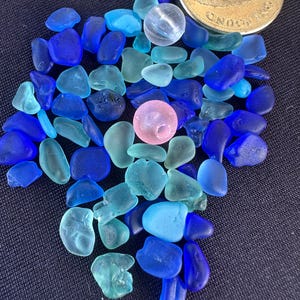 May include: A collection of small, smooth, tumbled sea glass pieces in various shades of blue and turquoise, mixed with one clear spherical bead and one pink spherical bead, positioned next to a gold-coloured one-pound coin for scale.