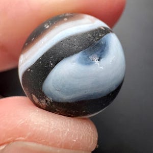 Peut inclure: Une bille vintage avec un motif tourbillonnant noir, blanc et bleu clair. La sphère de verre a une surface légèrement texturée et est tenue entre deux doigts. Un jouet et objet de collection classique.