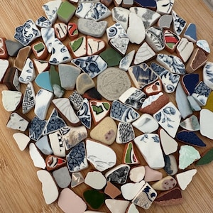 May include: A collection of broken ceramic pieces in various shapes and sizes, arranged on a wooden surface. The fragments display a range of colours, including white, blue, green, and brown, with floral and geometric patterns. A coin is also present.