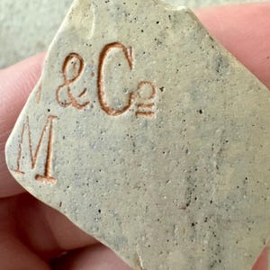 May include: A piece of pottery with the embossed text "& Co" and the letter "M" in a reddish-brown colour. The pottery has a speckled, light grey surface.