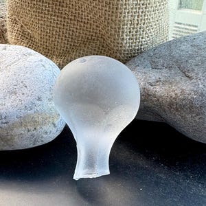 May include: A frosted glass bottle stopper with a bulbous top and a slender stem. The stopper is surrounded by grey stones and a hessian sack in the background. The stopper is a light, translucent colour.