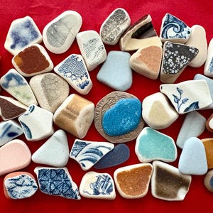 May include: A selection of sea pottery fragments on a red surface. The pieces vary in shape and size, displaying a palette of colours such as white, blue, brown, and beige, with floral and geometric designs.