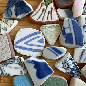 May include: A selection of ceramic fragments with diverse patterns and colours. The shards display blue and white floral, striped, and abstract designs, alongside pieces in green, pink, and brown hues. The fragments are arranged on a light brown surface.