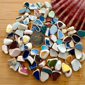 May include: A collection of sea pottery shards in various shapes and colours, including white, blue, brown, and yellow. A five pence coin is included for scale, and a section of a red and white seashell is visible.