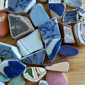 May include: A selection of sea pottery fragments in diverse shapes and sizes. The shards display blue and white patterns, solid blue, and other hues such as pink, brown, and green. The pieces are arranged on a light brown wooden surface.