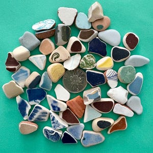 May include: A collection of sea glass pieces in various shapes and colors, including blue, white, brown, and green. Some pieces have decorative patterns. A coin is included for size reference. The sea glass is arranged on a turquoise background.