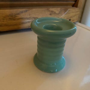 May include: A teal ceramic spool-shaped object with a circular opening at the top. The object has a glossy finish and a ribbed design on the body. It sits on a white surface.