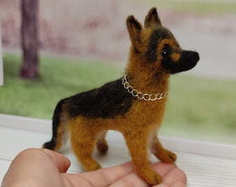 May include: A small, hand-felted German Shepherd dog figurine with black and brown fur. The figurine is wearing a silver chain collar and is standing on a person's hand.