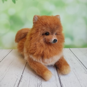 Puede incluir: Figura hecha a mano de un perro Pomerania naranja con un pelaje esponjoso, tumbado sobre una superficie de madera blanca. El perro tiene una nariz negra y ojos pequeños y oscuros. El fondo es verde suave con hojas borrosas.