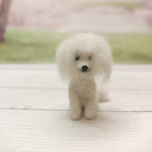 May include: A white needle felted poodle dog figurine. The dog is sitting on a white wooden surface.