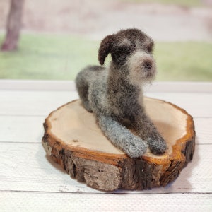 May include: A needle felted German Shorthaired Pointer dog figurine, lying down on a wooden slice. The dog is gray and brown with white markings.