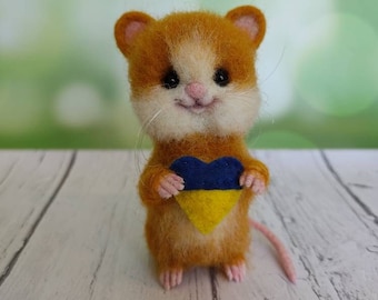 Può includere: Un piccolo topo di feltro arancione e bianco che tiene un cuore giallo con un design blu e giallo, che rappresenta la bandiera ucraina.