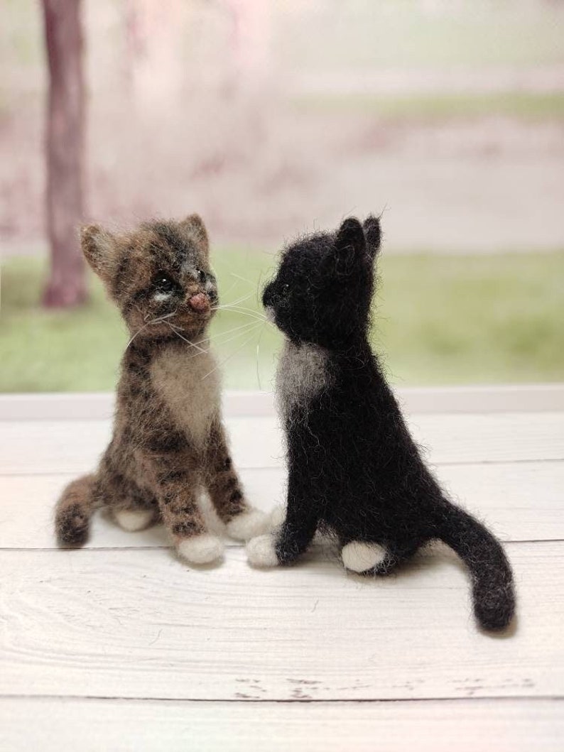 May include: Two small felted cat figurines, one brown tabby and one black with white markings, sitting on a white wooden surface.