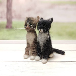 May include: Two felted wool kittens, one brown tabby and one black with white markings, sitting side by side on a white wooden surface.