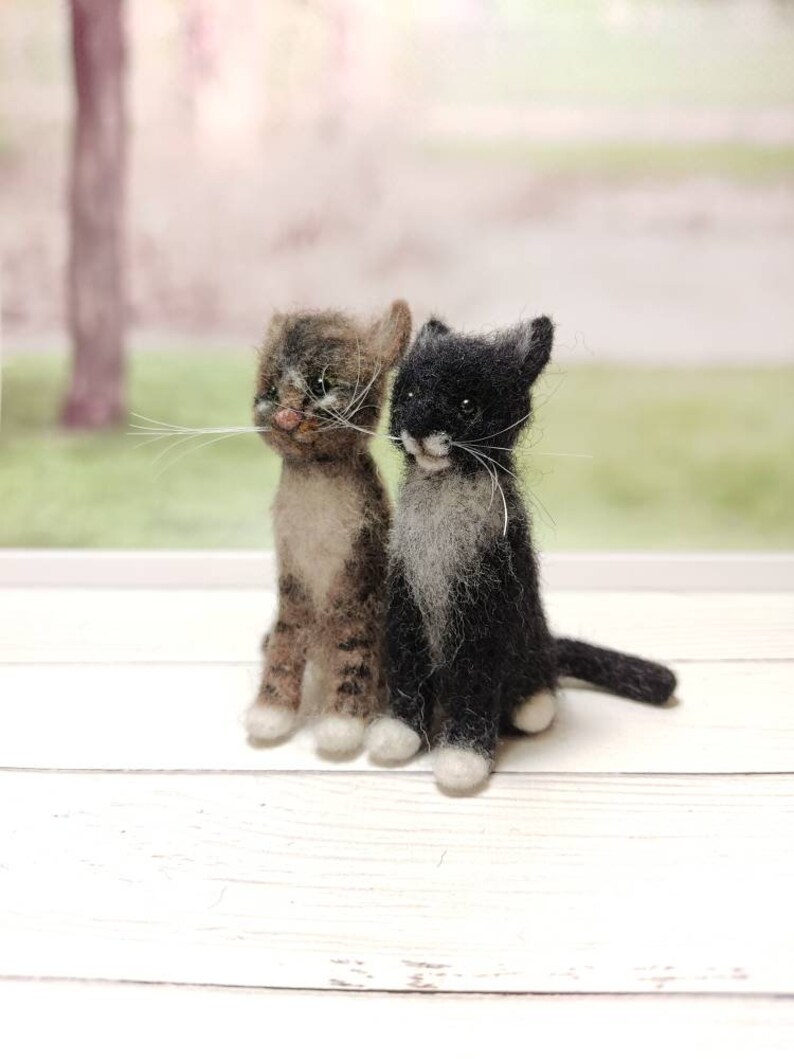 May include: Two felted wool kittens, one brown tabby and one black with white markings, sitting side by side on a white wooden surface.
