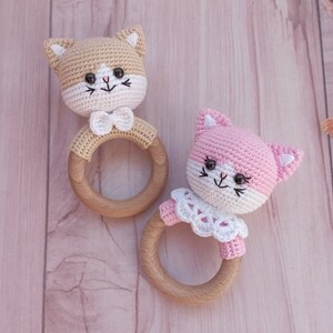 May include: Two crocheted cat rattle toys with wooden rings. One cat is tan with a white bow tie and the other is pink with a white lace collar.