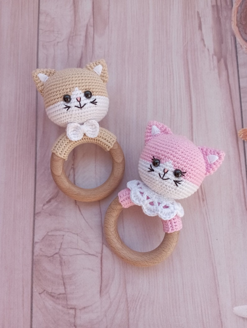 May include: Two crocheted cat rattle toys with wooden rings. One cat is tan with a white bow tie and the other is pink with a white lace collar.