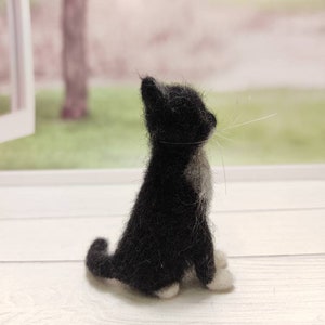 May include: A black and white needle felted cat figurine sitting on a white wooden surface. The cat is looking up and has a white patch on its chest.