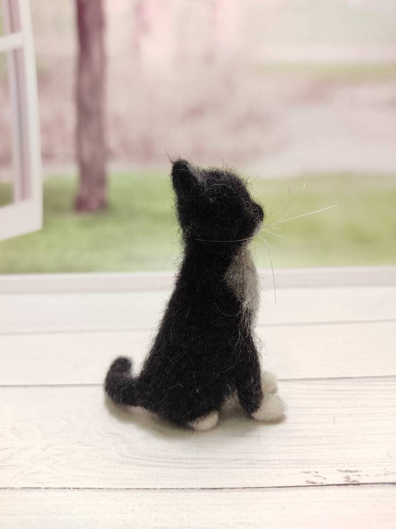 May include: A black and white needle felted cat figurine sitting on a white wooden surface. The cat is looking up and has a white patch on its chest.