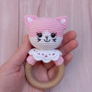 May include: Pink crocheted cat rattle with a white crocheted collar and a wooden ring.