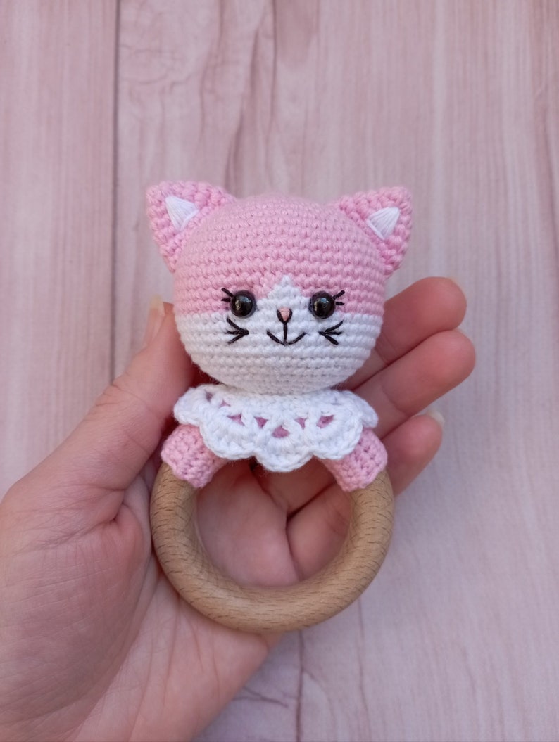 May include: Pink crocheted cat rattle with a white crocheted collar and a wooden ring.