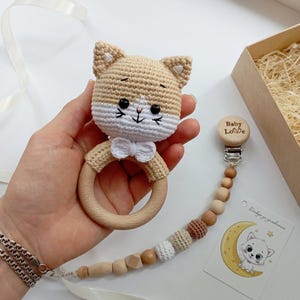 May include: A crocheted beige cat rattle with a white bow and wooden ring. The rattle is attached to a wooden clip with the words "Baby Love" engraved on it. The clip is connected to a chain of wooden and crocheted beads.