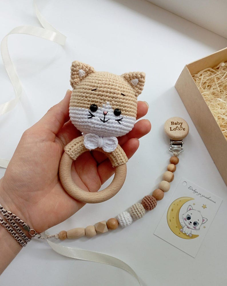 May include: A crocheted beige cat rattle with a white bow and wooden ring. The rattle is attached to a wooden clip with the words "Baby Love" engraved on it. The clip is connected to a chain of wooden and crocheted beads.