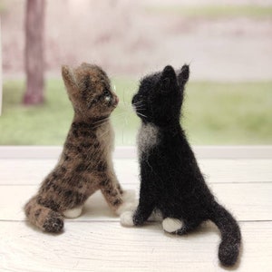 May include: Two felted wool kittens, one brown tabby and one black and white, are sitting facing each other on a white wooden surface.