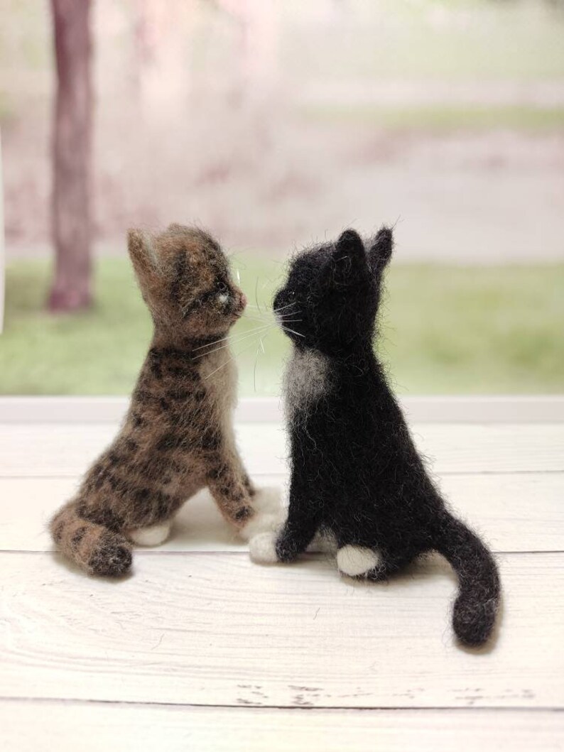 May include: Two felted wool kittens, one brown tabby and one black and white, are sitting facing each other on a white wooden surface.
