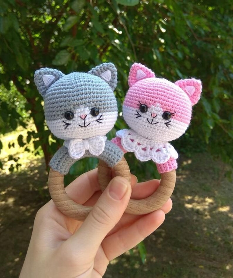 May include: Two crocheted cat rattle toys with wooden rings. One cat is gray and the other is pink. Both cats have white faces and white bows.