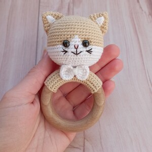 May include: A crocheted cat rattle toy with a wooden ring. The cat is beige with white accents and black eyes. The toy has a white bow.