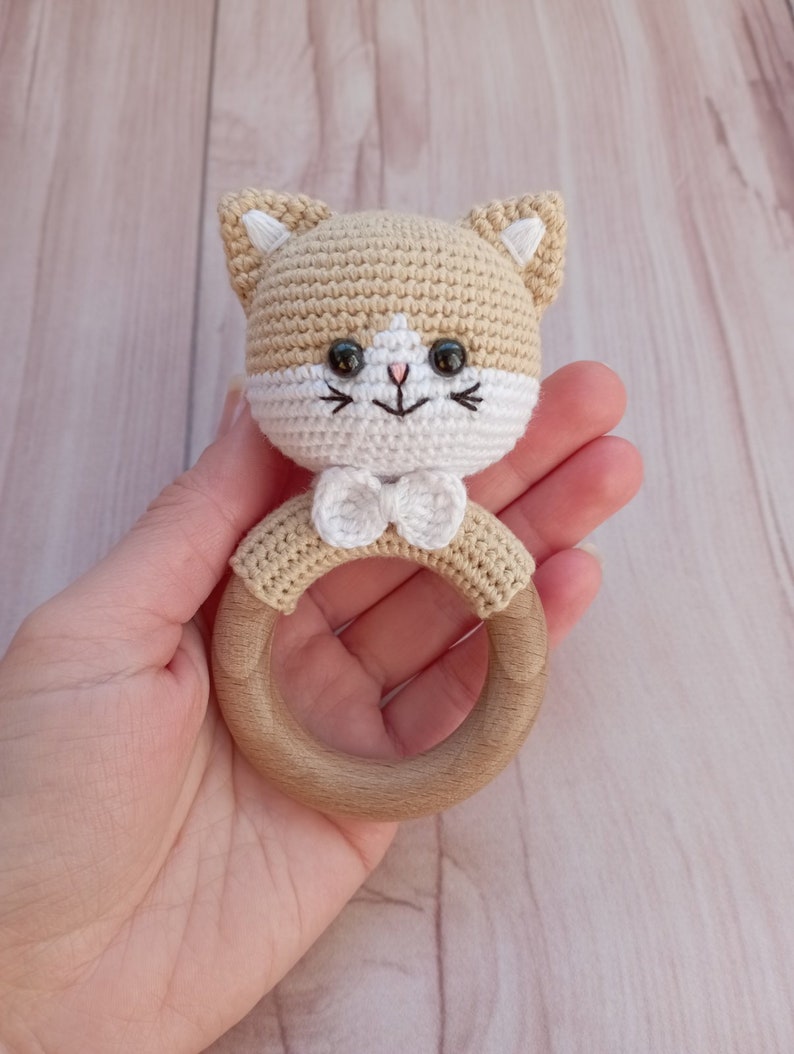 May include: A crocheted cat rattle toy with a wooden ring. The cat is beige with white accents and black eyes. The toy has a white bow.