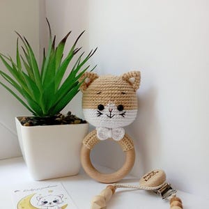 May include: A crocheted cat rattle toy with a wooden ring and a white bow. The toy is beige and white with black eyes and a pink nose. The toy is attached to a wooden clip with the text "Baby Love" on it.