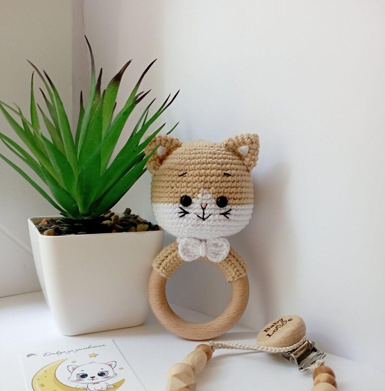 May include: A crocheted cat rattle toy with a wooden ring and a white bow. The toy is beige and white with black eyes and a pink nose. The toy is attached to a wooden clip with the text "Baby Love" on it.