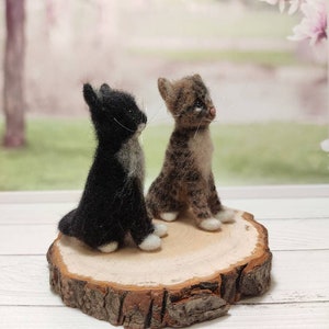 May include: Two felted wool cat figurines, one black and white and one brown and white, sitting on a wooden slice.