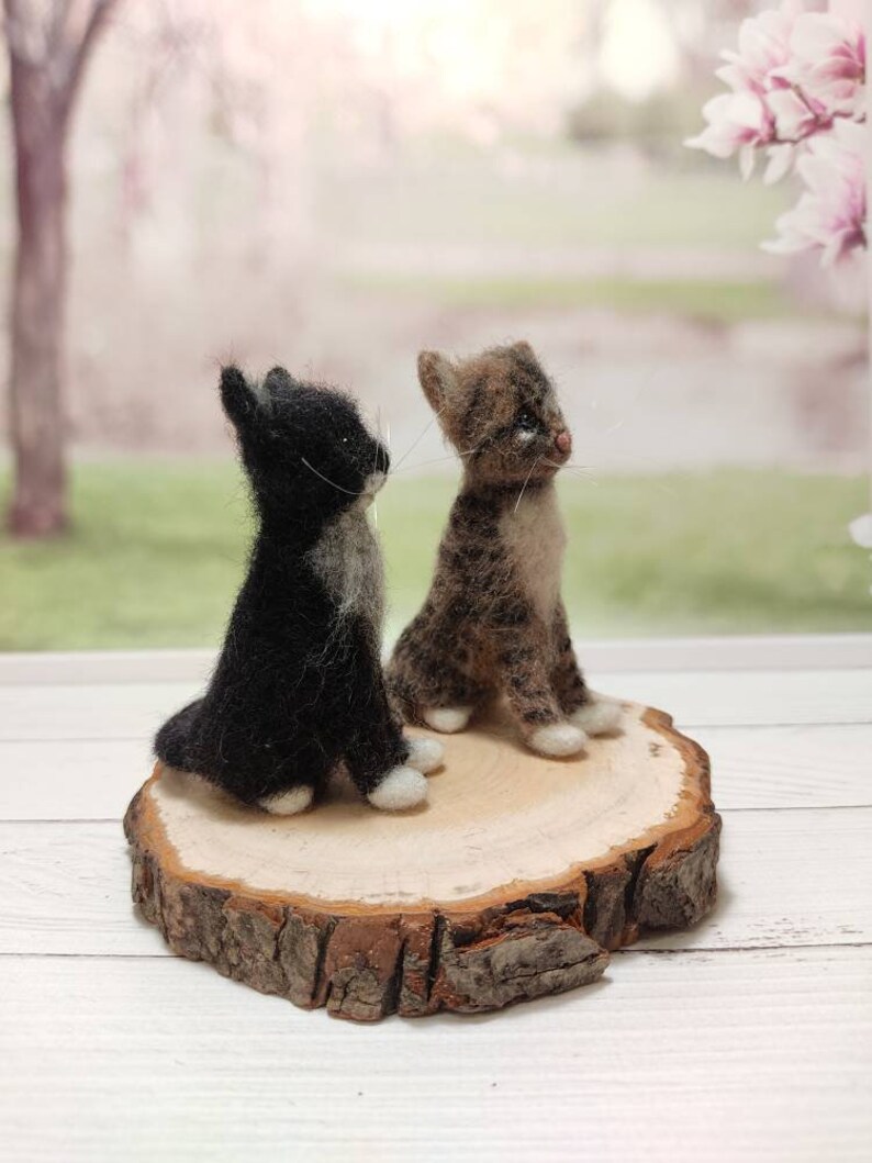 May include: Two felted wool cat figurines, one black and white and one brown and white, sitting on a wooden slice.