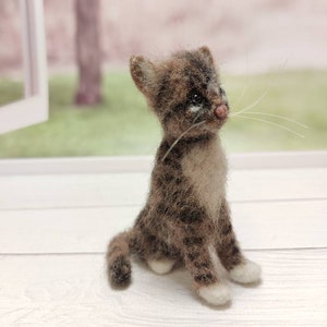 May include: A small, brown and white needle felted cat figurine with white paws and a white chest. The cat is sitting on a white wooden surface.