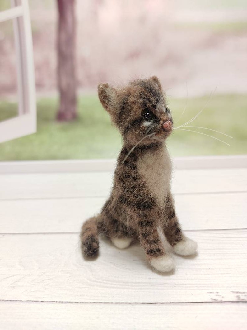 May include: A small, brown and white needle felted cat figurine with white paws and a white chest. The cat is sitting on a white wooden surface.