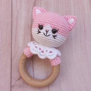 May include: A pink and white crocheted cat rattle with a wooden ring. The cat has black eyes and a white face with a pink nose.