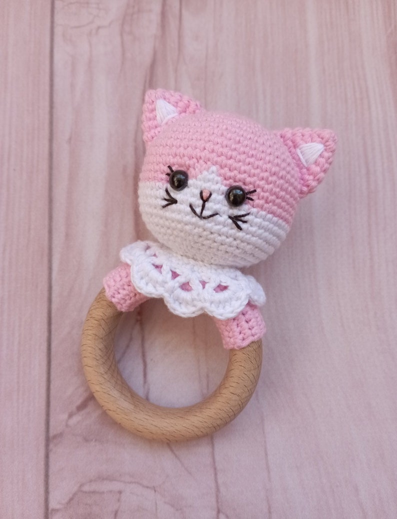 May include: A pink and white crocheted cat rattle with a wooden ring. The cat has black eyes and a white face with a pink nose.