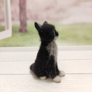 May include: A small black and white needle felted cat figurine sitting on a white wooden surface. The cat is looking to the right and has white paws and a white patch on its chest.