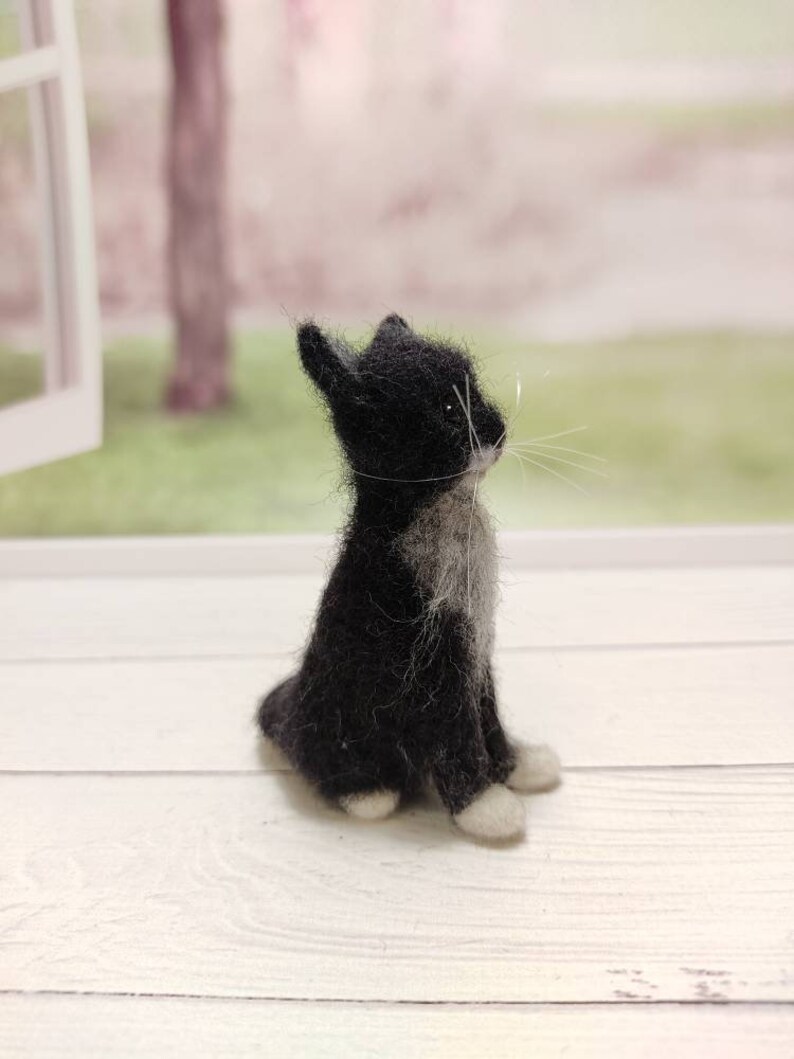 May include: A small black and white needle felted cat figurine sitting on a white wooden surface. The cat is looking to the right and has white paws and a white patch on its chest.