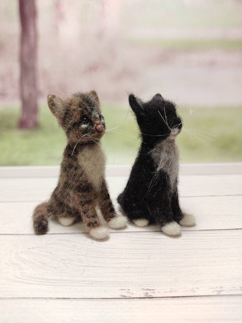 May include: Two felted wool cat figurines, one brown tabby with white markings and one black with white markings, sitting on a white wooden surface.