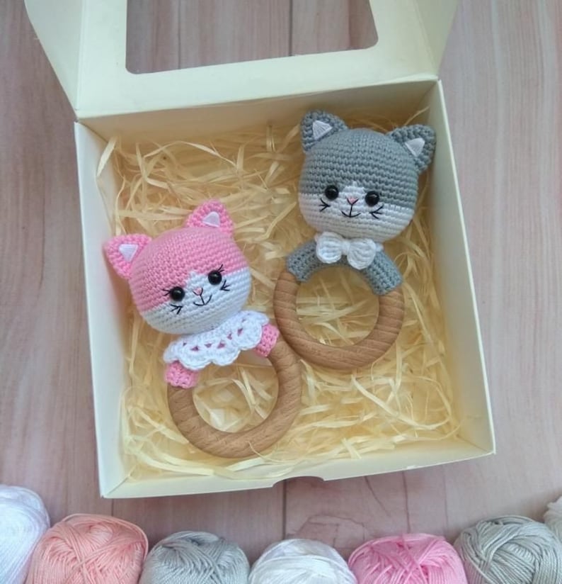 May include: Two crocheted cat rattle toys in a white box with wood rings. The cat on the left is pink with white accents and the cat on the right is gray with white accents.