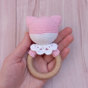 May include: Pink and white crocheted baby rattle with a wooden ring. The rattle has a crocheted animal head with ears and a white crocheted collar.