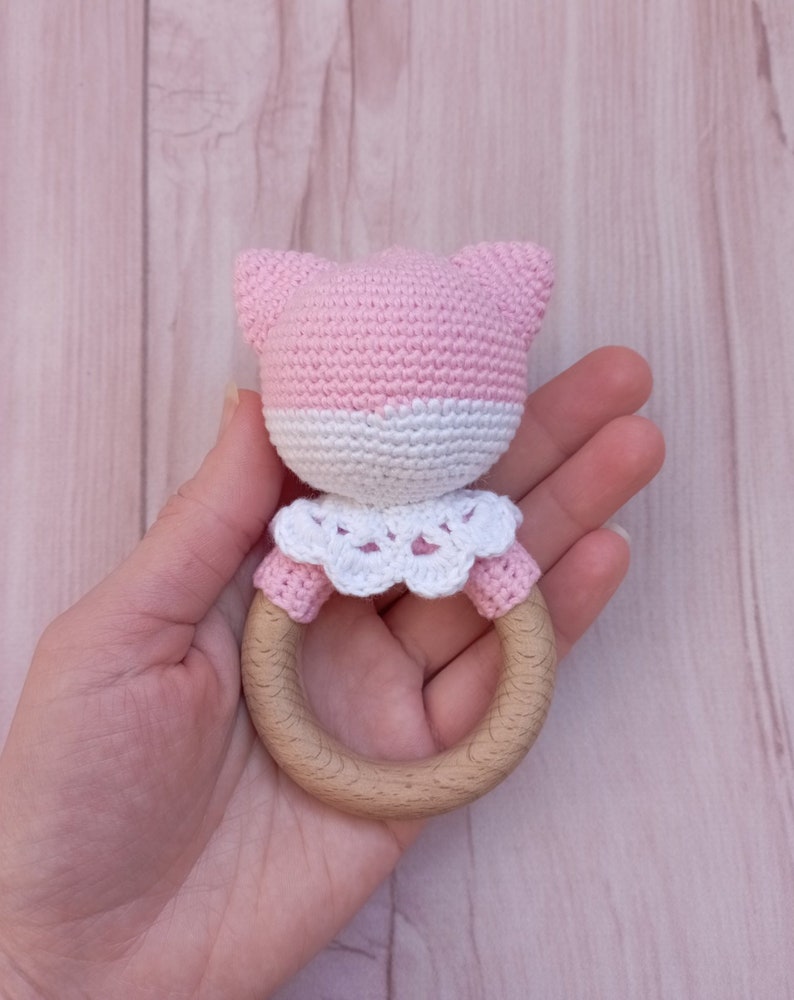 May include: Pink and white crocheted baby rattle with a wooden ring. The rattle has a crocheted animal head with ears and a white crocheted collar.