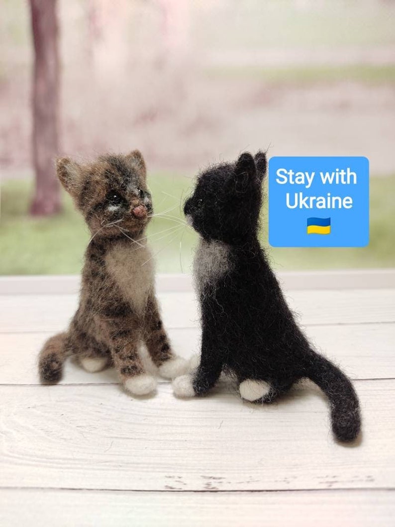 May include: Two needle felted cat figurines, one brown tabby and one black and white, sitting on a white wooden surface. The text "Stay with Ukraine" is in a blue box with the Ukrainian flag.